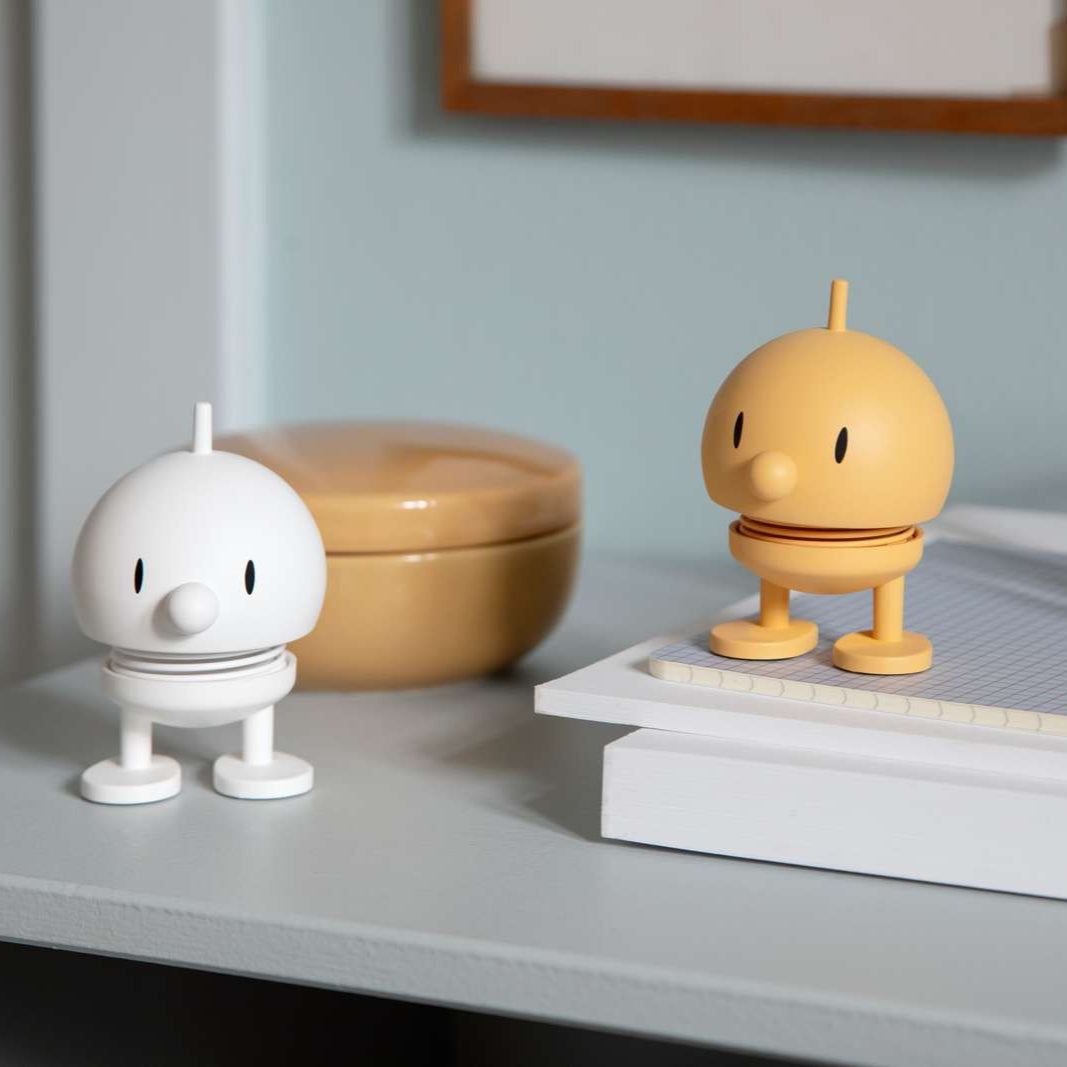 Two small figurines, one white and one yellow, on a surface with a light blue wall in the background.