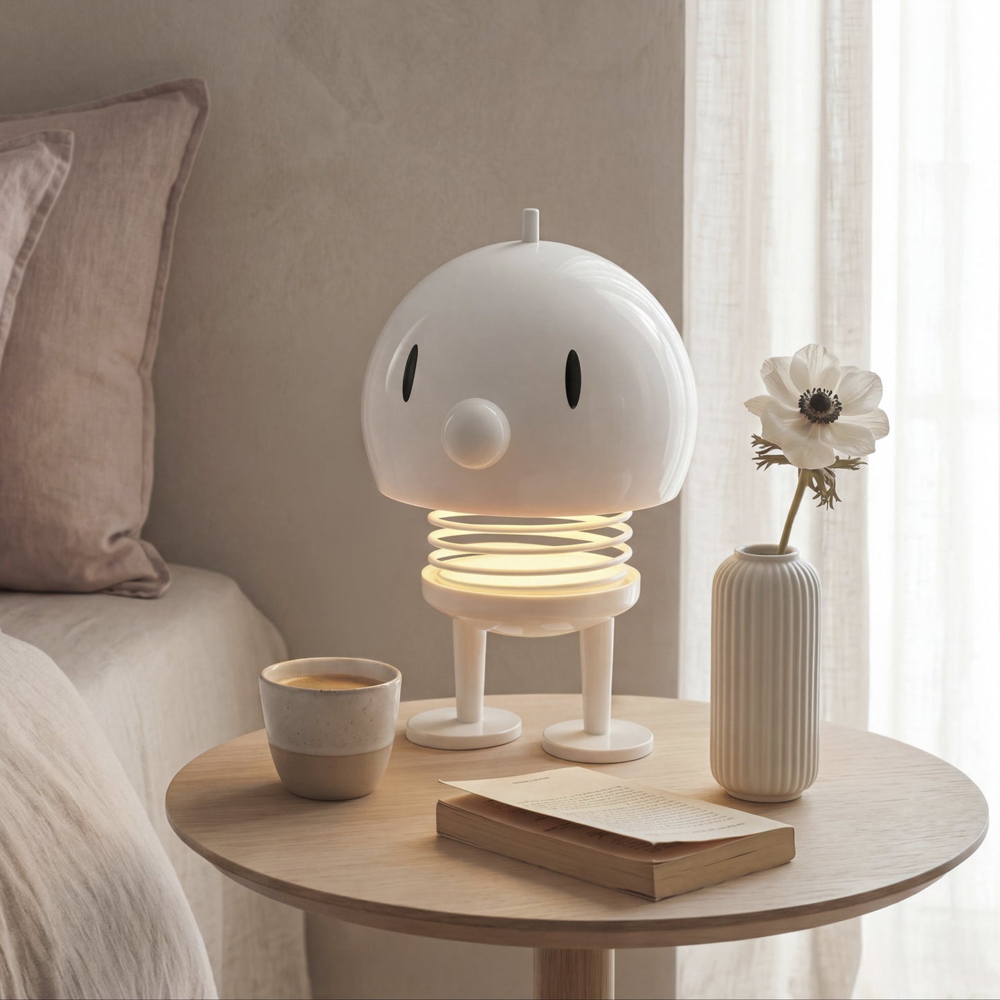 A whimsical white robot-shaped night light with a glowing spring base sits on a round wooden bedside table next to a bed. The scene is styled with a small ceramic cup, a book, and a ribbed vase holding a dried flower, all set against a soft beige wall and white sheer curtains.