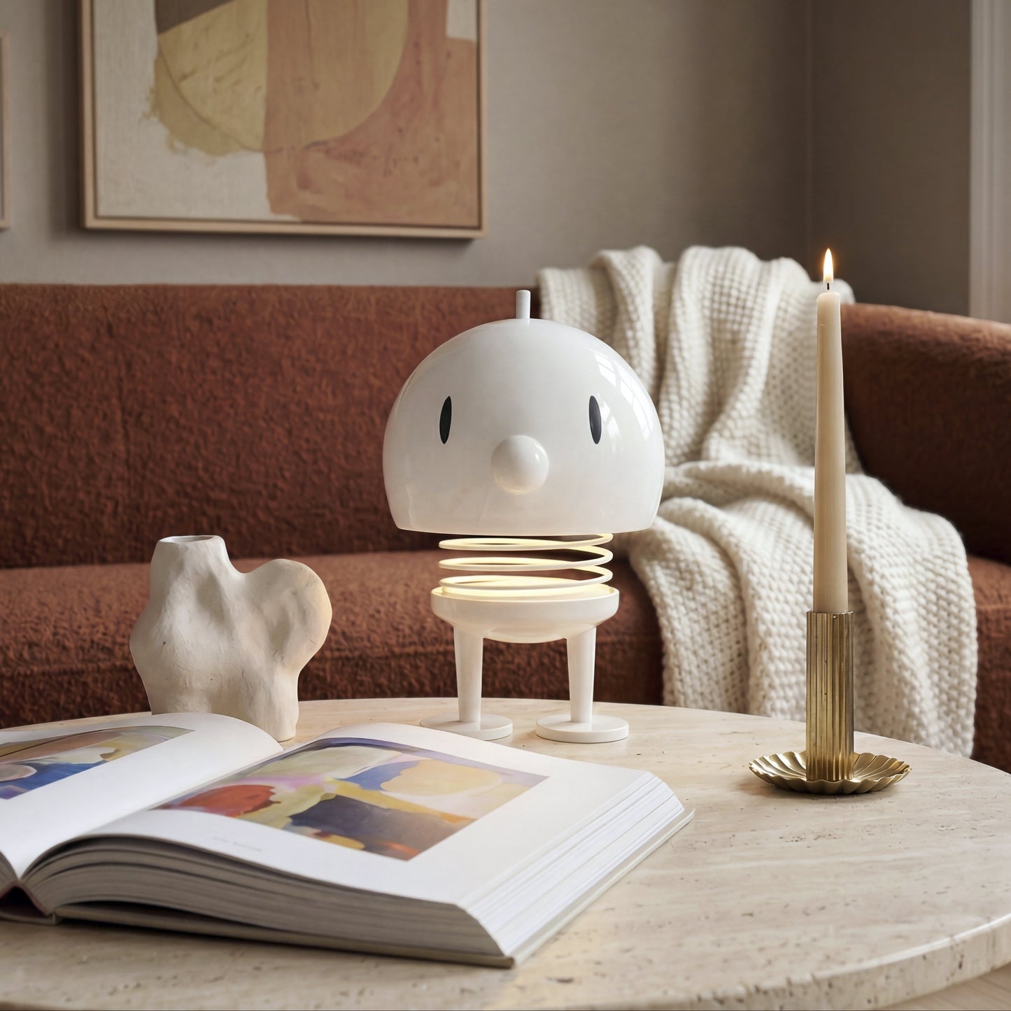 A white, minimalist robot-shaped lamp with a spring mechanism stands on a round stone coffee table in a cozy living room setting. The scene includes an open book, a small white sculpture, a gold candlestick holder with a lit candle, and a brown textured sofa in the background.