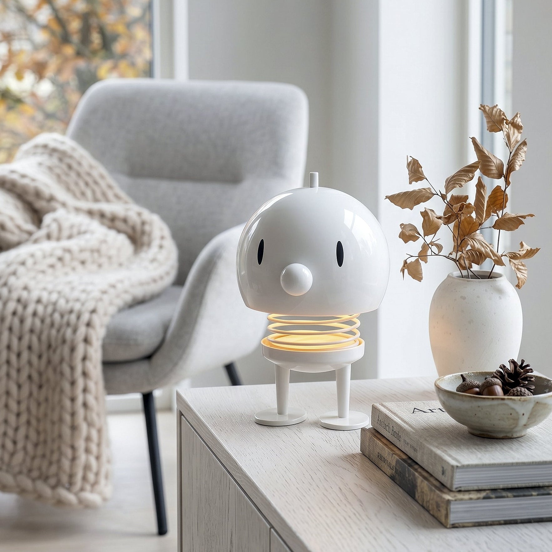 A small white cartoon-style lamp with a glowing warm yellow spring base sits on a light wood console table in a bright living room. A gray armchair with a cream knit throw and a small vase with dried leaves are nearby.