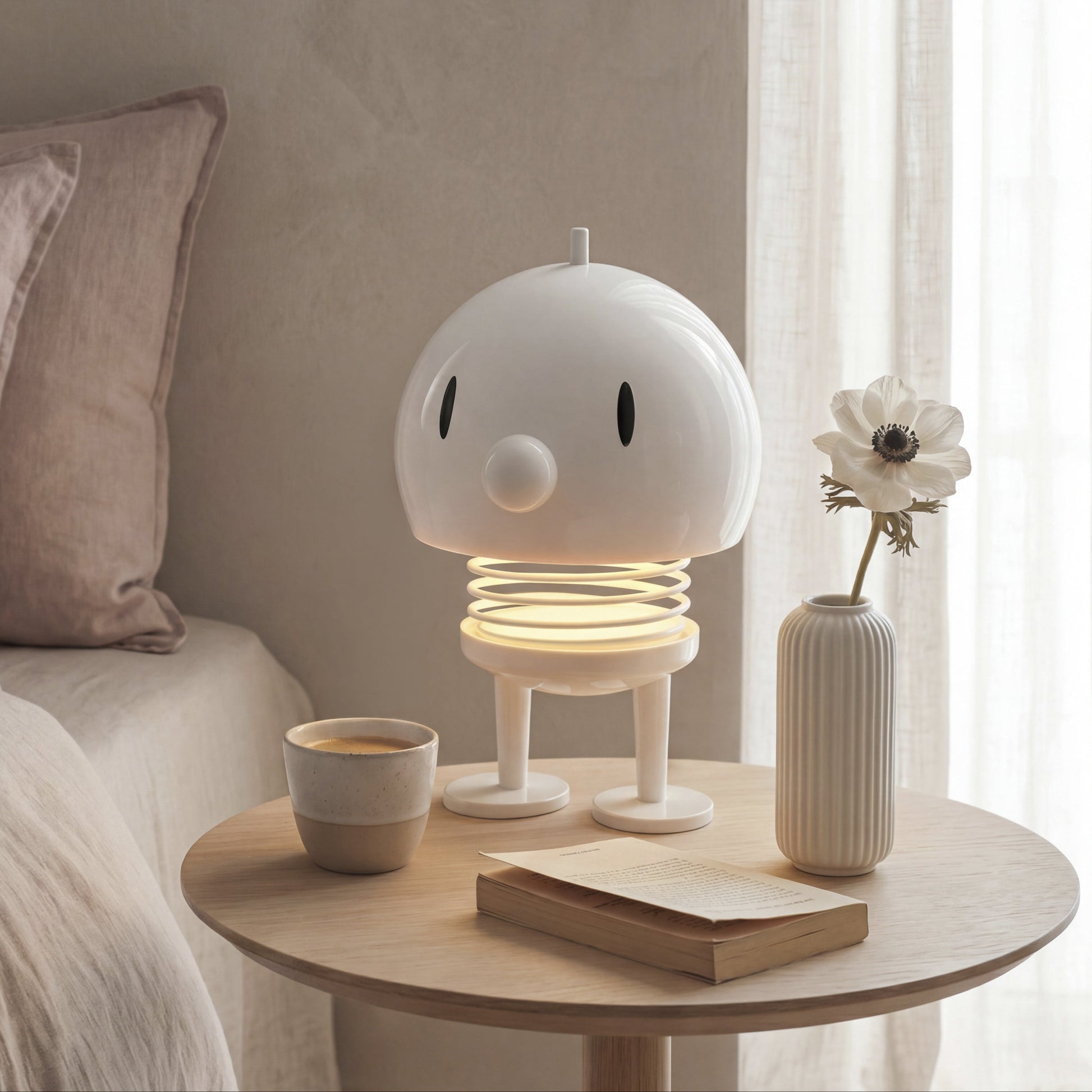 A whimsical white robot-shaped night light with a glowing spring base sits on a round wooden bedside table next to a bed. The scene is styled with a small ceramic cup, a book, and a ribbed vase holding a dried flower, all set against a soft beige wall and white sheer curtains.