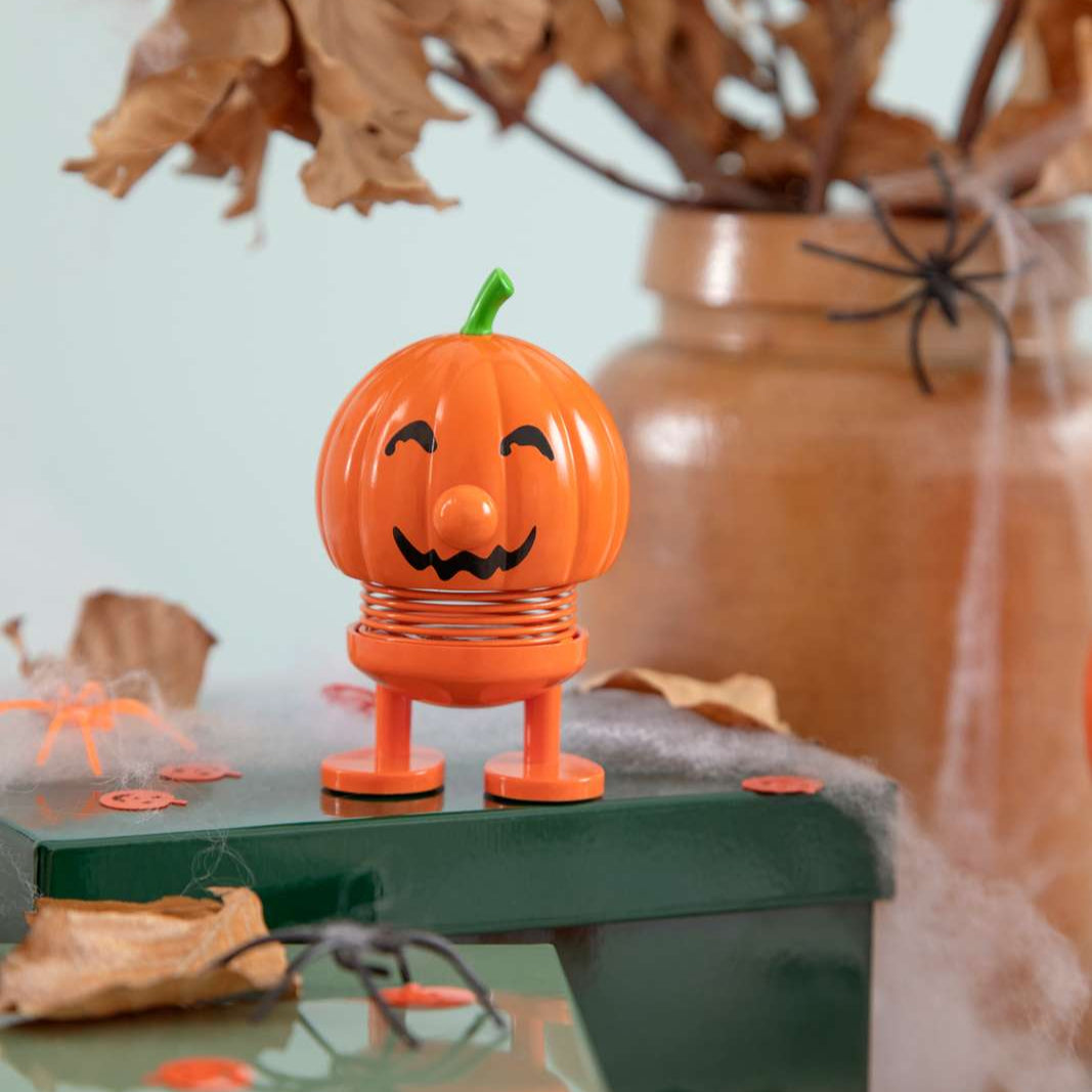 Orange jack-o'-lantern toy with a smiling face on a green surface with autumn leaves and spider decorations.