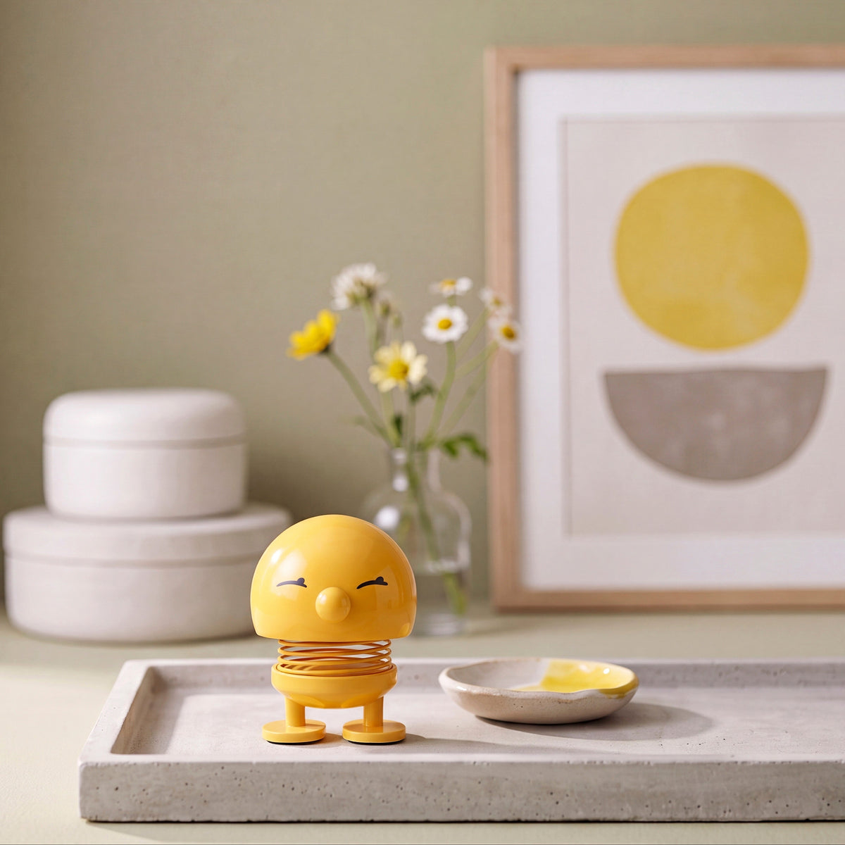 A small yellow spring toy figure with a sleeping face sits on a light gray tray next to a shallow dish with yellow wax, styled in a warm, minimalist room with a framed art print and flowers in the background.
